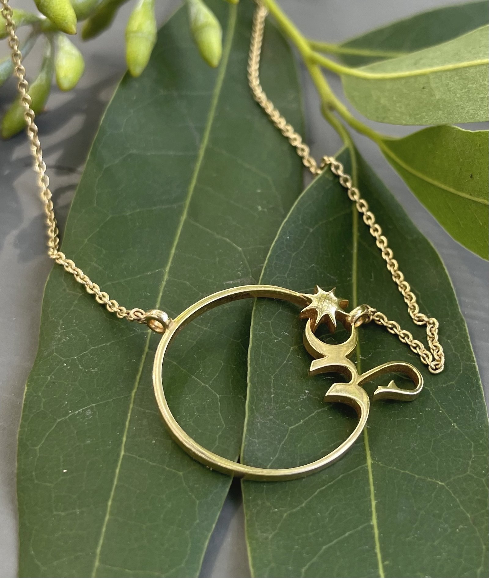 Gold Om pendant necklace resting on eucalyptus leaves